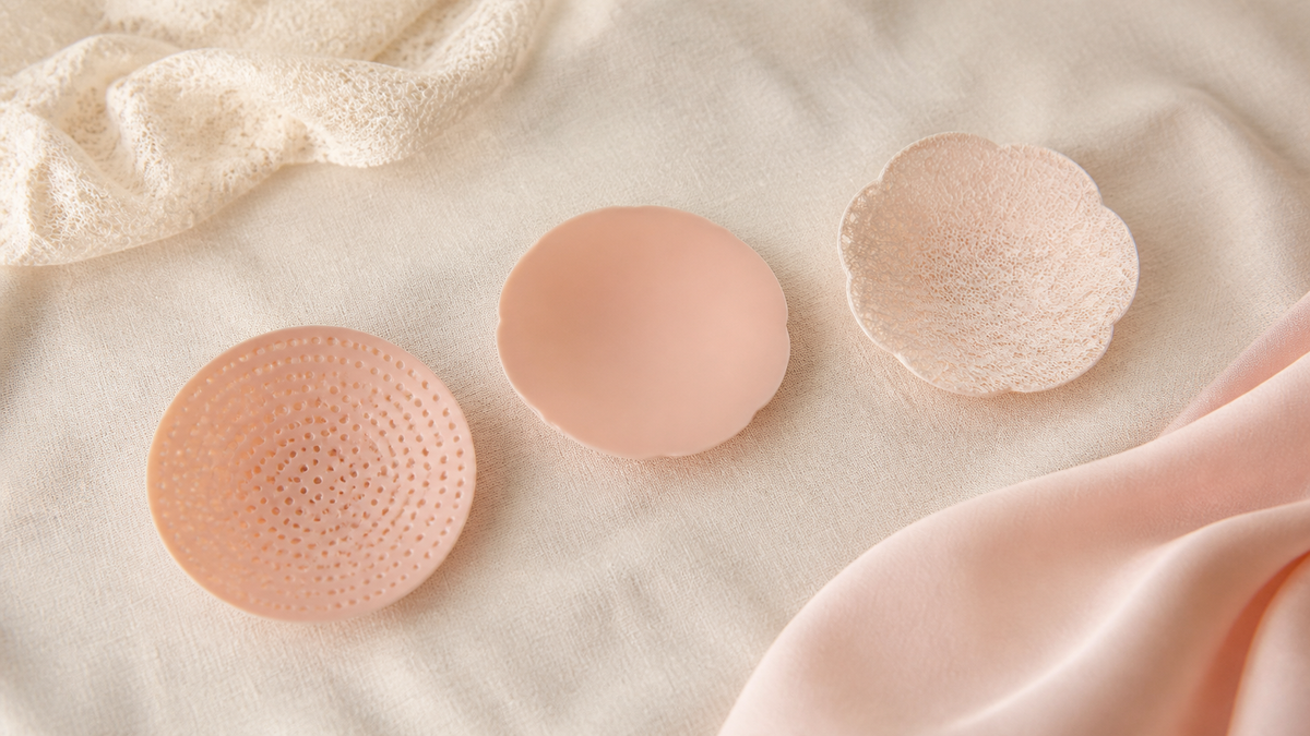 Three nipple covers laid apart on cream linen: a perforated silicone disc, a smooth silicone disc and a fine lace petal cover