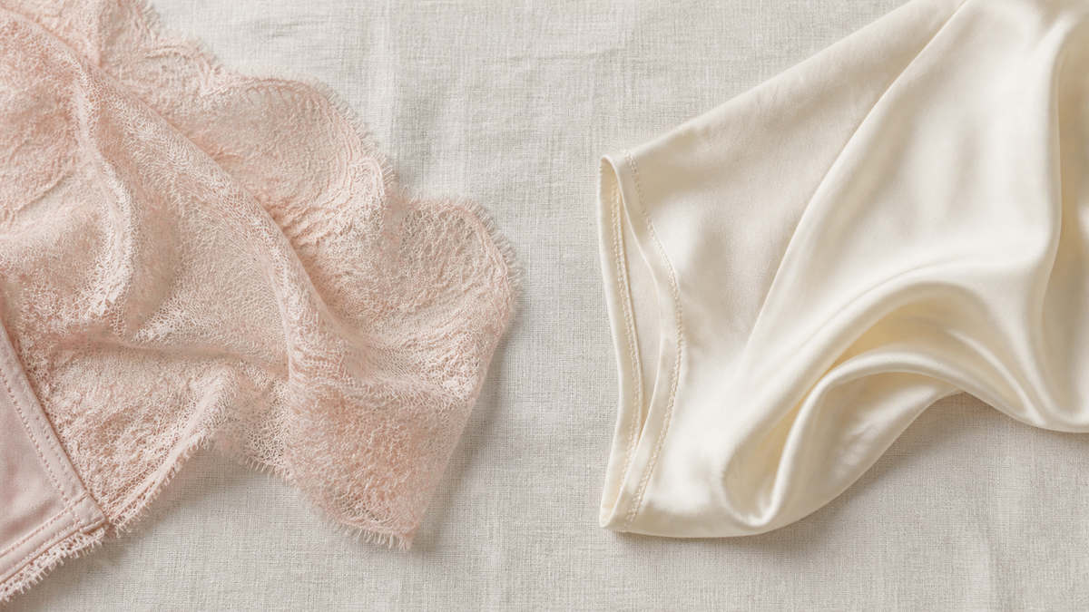 Flat lay on cream linen of a blush French lace lingerie panel beside a smooth ivory silk panel, showing the difference in weight and opacity