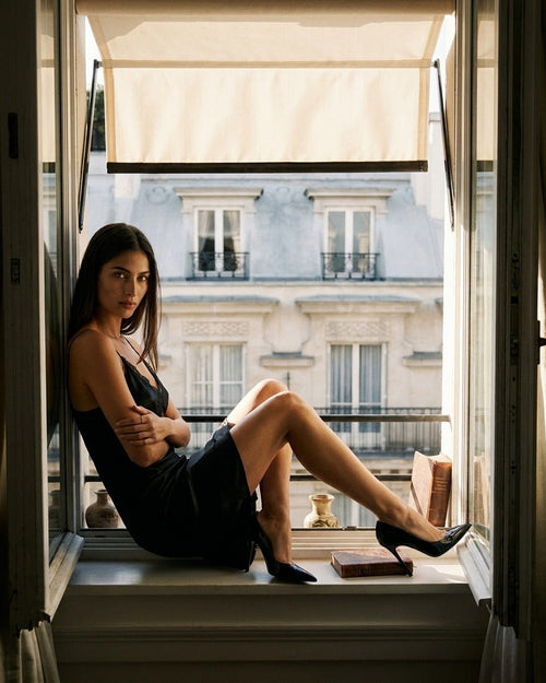 Darette Backhanded Silk Slip in Noir - sitting on an open window ledge in the black silk slip with lace trim, buildings across the street behind
