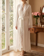 Darette Late Start Robe in Ivory - long satin robe with lace cuffs, front view beside a garden window