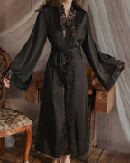 Darette Late Start Robe in Noir - long satin robe with lace cuffs, standing beside a chair