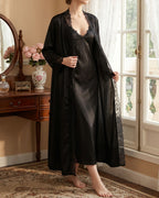 Darette Late Start Robe in Noir - long satin robe with lace trim, standing beside a vanity mirror