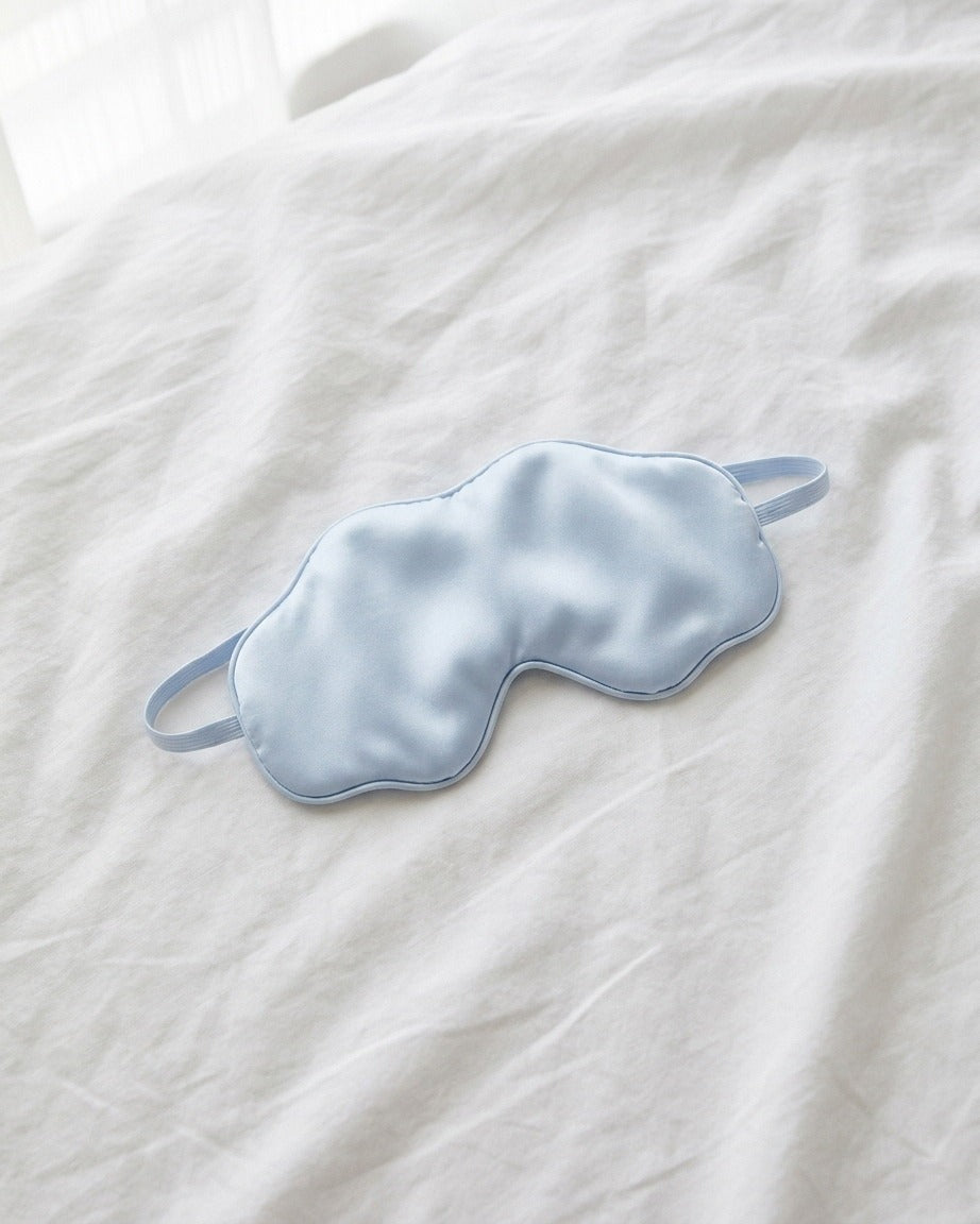 Darette Lights Out Silk Eye Mask in Sky laid flat on white bedding, showing the elastic strap