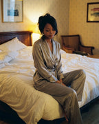 Darette The Long Weekend Silk Set in Mist - shirt and trousers worn seated on the edge of a hotel bed in lamplight